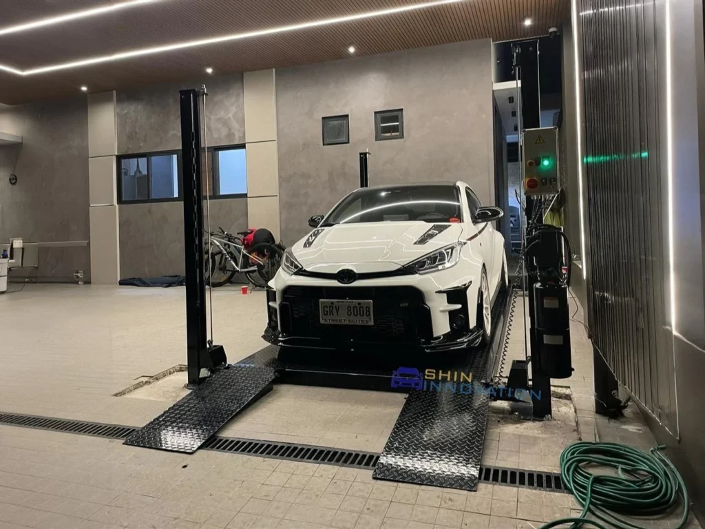 Four post-style car lift in a modern concrete garage, with a white Toyota GR Yaris positioned on the platform.