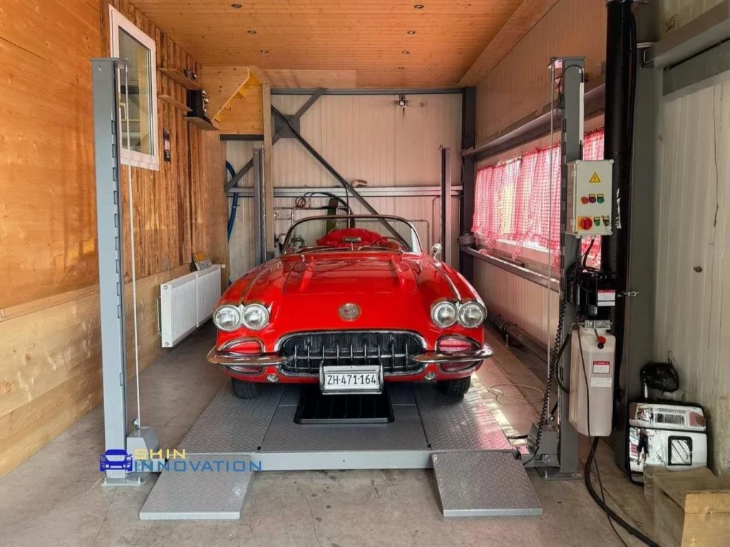 Four-post car lift in a wooden garage, lifting a classic red Chevrolet Corvette.